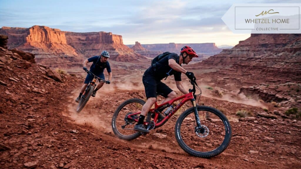 A photorealistic action shot of two mountain bikers riding a red dirt trail at dawn. Soft, cool morning light illuminating the dramatic red rock formations in the background. Minimalist gear, athletic lifestyle, high resolution, vivid contrasting colors.