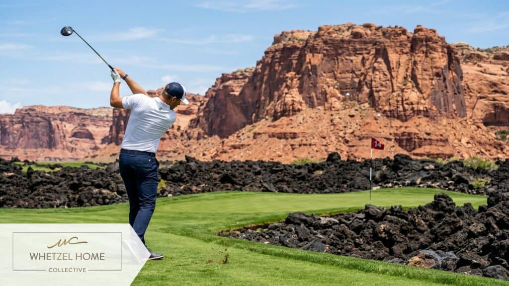 A male golfer mid-swing on a vibrant emerald green fairway. The golf hole is dramatically carved out of rugged black basalt lava rock formations. In the immediate background, massive and striking red Navajo sandstone cliffs rise steeply under a bright midday sun. High shutter speed, photorealistic, sports photography style, rich contrasting colors.