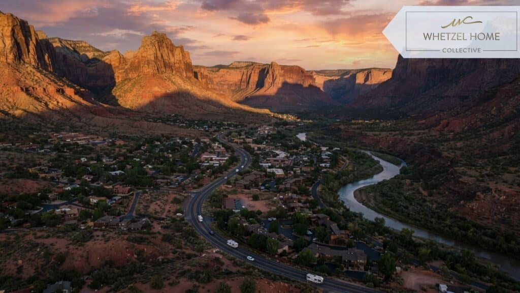 An aerial view of a winding desert road passing through a small rural town near a national park, red soil, sparse green desert vegetation, the Virgin River snaking through the valley, dramatic shadows from the setting sun, cinematic photography.